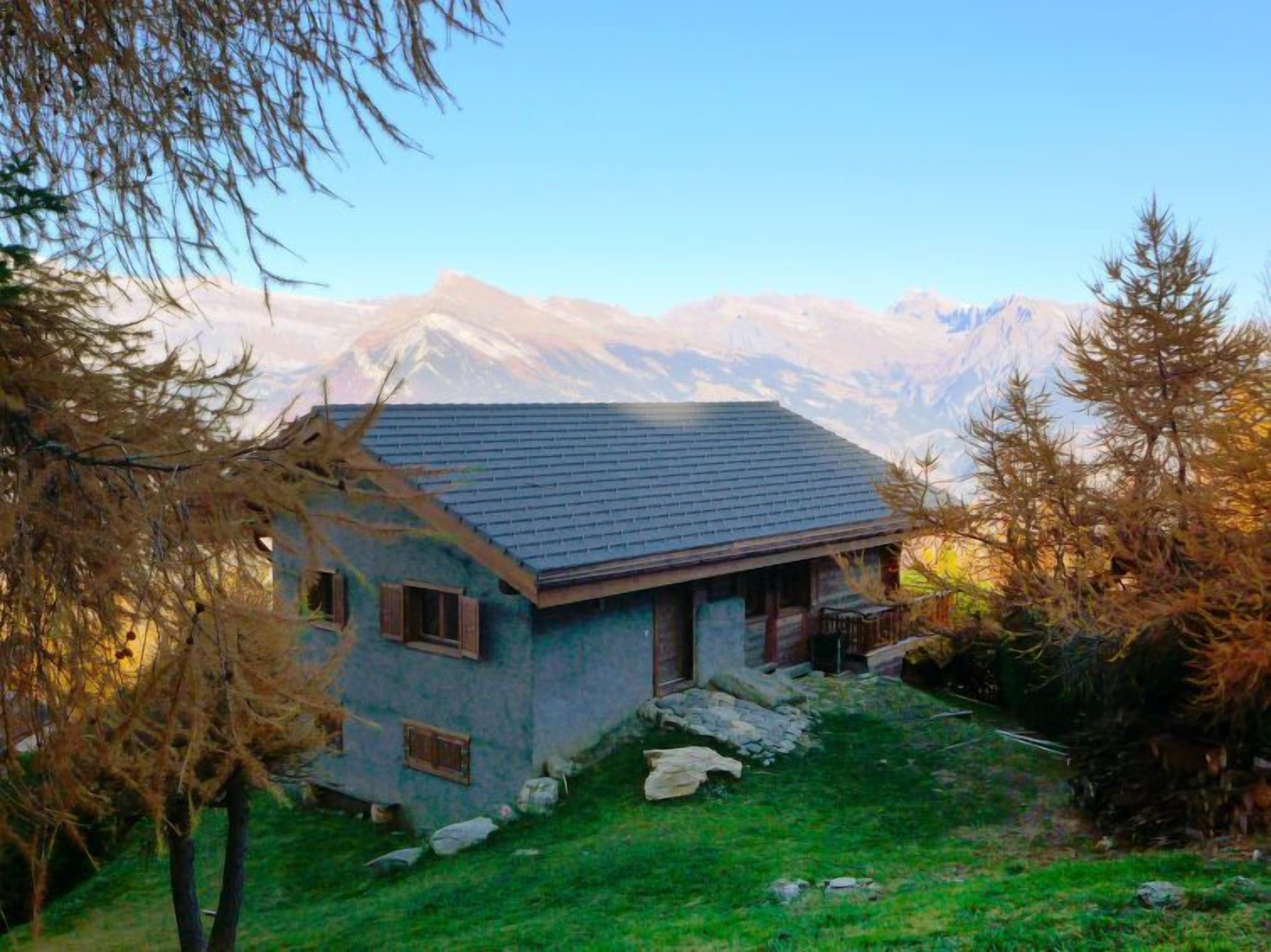 A small house with a sloped roof, surrounded by a grassy area and trees, with a mountain range in the background.