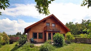 A wooden cabin with a balcony, surrounded by a lush garden and a wooden fence.