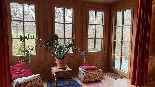 A cozy room with wooden walls and large windows, featuring wicker furniture and a potted plant.