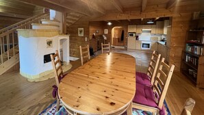 A spacious wooden interior with a large round table and chairs, a staircase, a kitchen area, and a bookshelf.