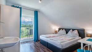 A bedroom with a bed, bedside table, lamp, and a view of greenery through a window.