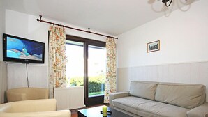 Ein Wohnzimmer mit einem beigen Sofa, einem schwarzen Couchtisch, einem beigen Sessel und einem Fernseher an der Wand.
