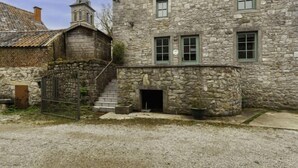 A stone building with a small staircase leading to a door, a church steeple in the background, and a gravel driveway. - La Bastide by Interhome (Thy-le-Bauduin)