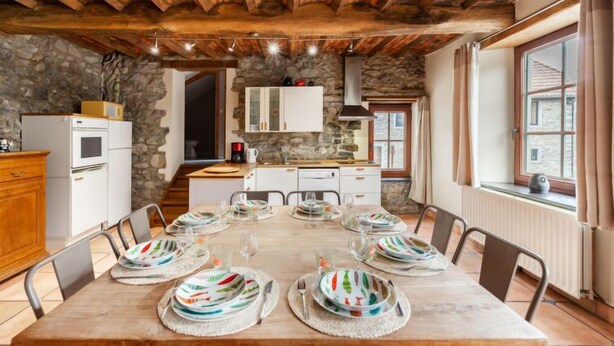A dining area with a wooden table set for six, stone walls, a kitchen with white cabinets, and a stone ceiling. - La Bastide by Interhome (Thy-le-Bauduin)