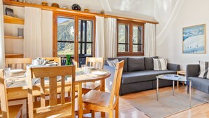 Ein gemütliches Wohnzimmer mit einem Holzesstisch, Stühlen, einer Couch und einem Couchtisch. Große Fenster bieten Blick auf eine Berglandschaft.