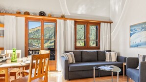 Ein gemütliches Wohnzimmer mit einer Couch, einem Glastisch, Holzstühlen und Blick auf eine Berglandschaft durch große Fenster.