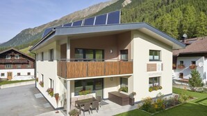 Un edificio moderno de dos pisos con balcón, paneles solares y un pequeño espacio al aire libre con asientos.