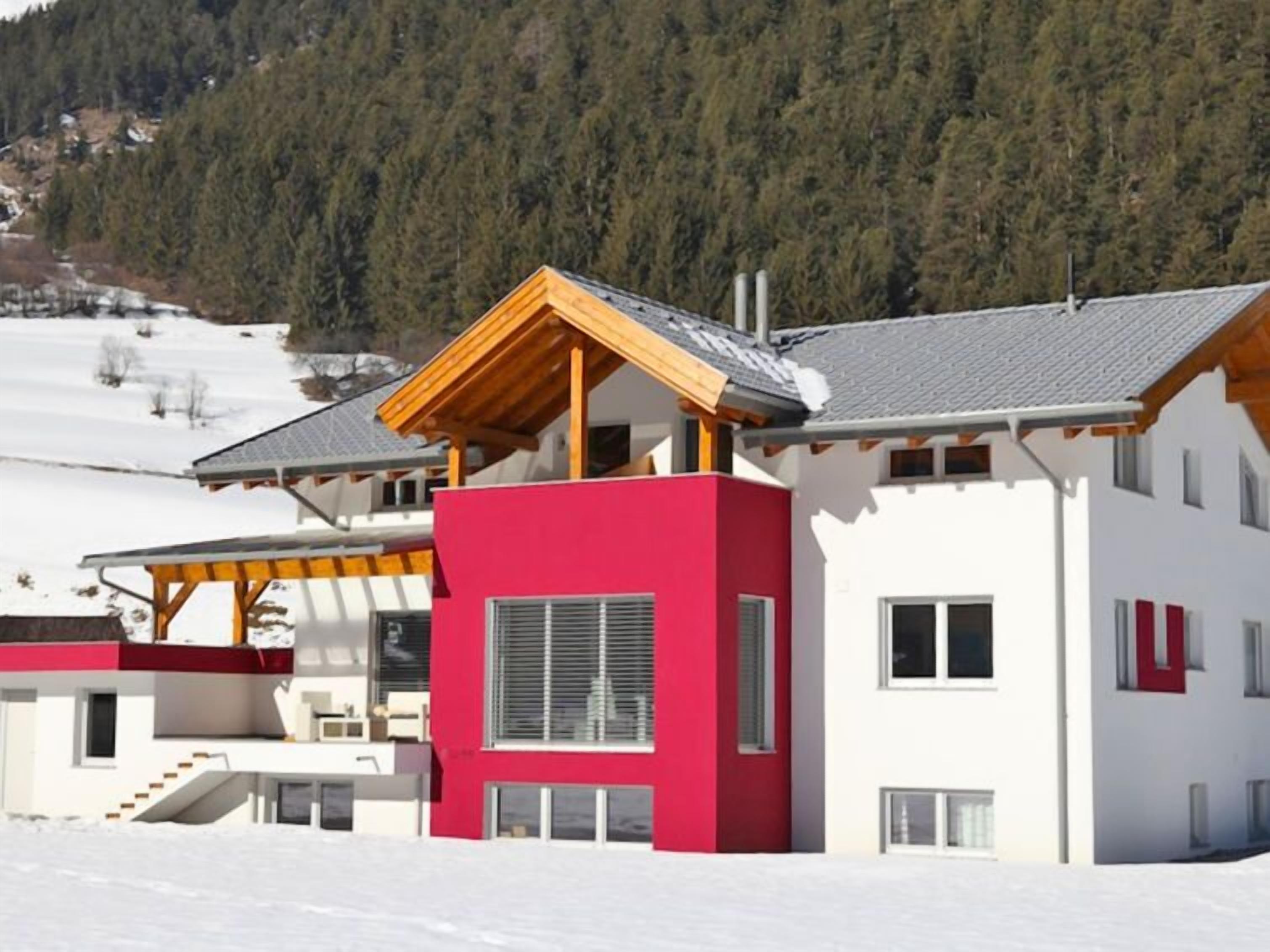 Un edificio moderno de dos pisos con una decoración en rojo y blanco, situado en un paisaje nevado con una colina boscosa al fondo.