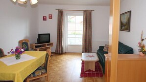 A cozy living room with a wooden floor, a dining table with a yellow tablecloth, a television on a stand, a sofa, and a window with curtains.