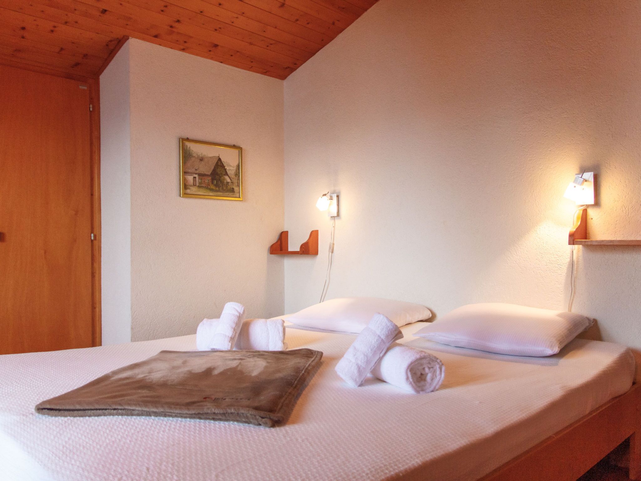 A neatly made bed with white linens and rolled towels, a painting on the wall, and two wall-mounted lamps.
