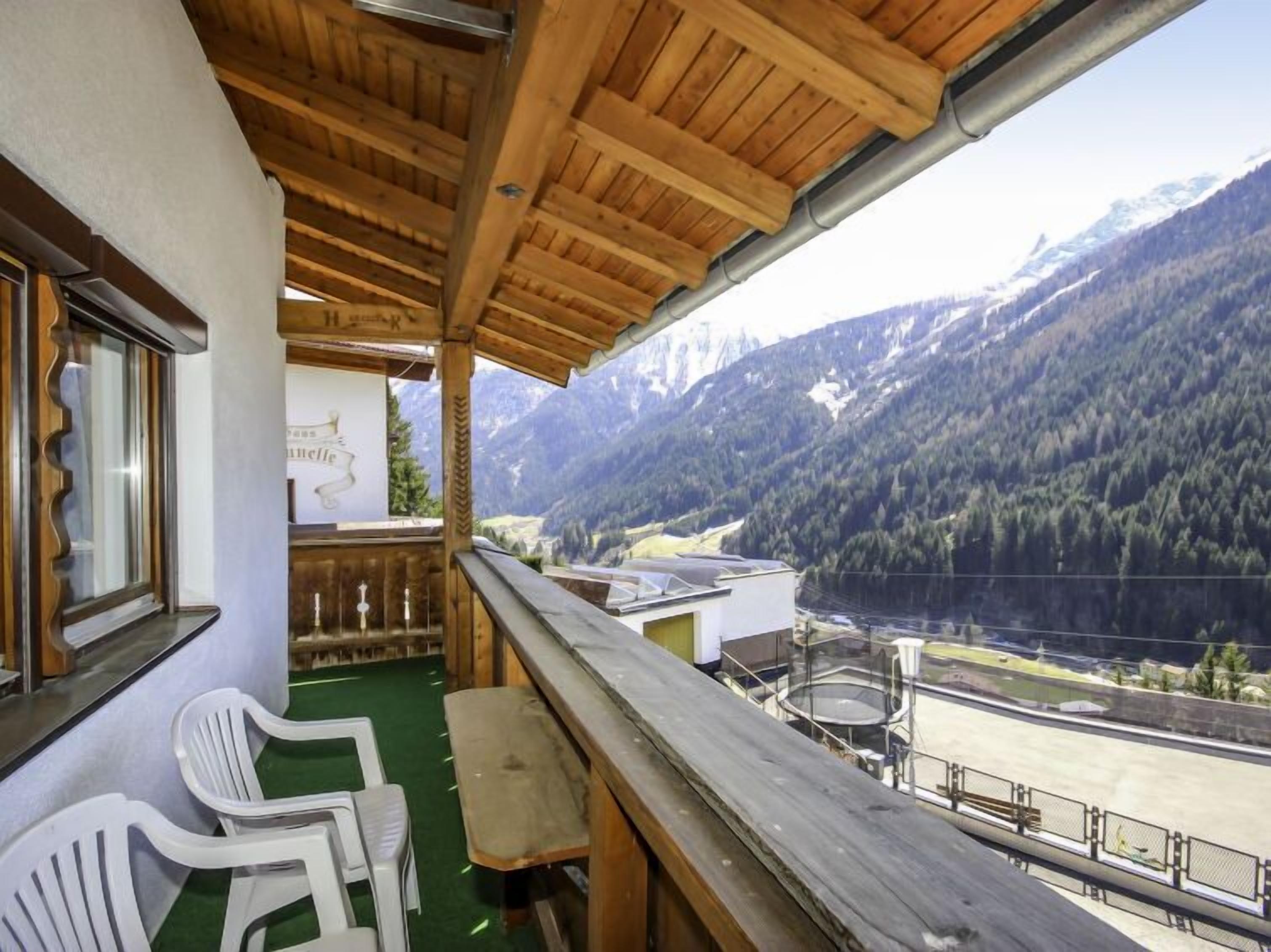 Ein Balkon mit Holzbalkonen, der auf ein Tal mit Bergen blickt. Auf dem Balkon befinden sich zwei weiße Plastikstühle und ein Tisch.