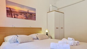 Una cama bien hecha con almohadas, una lámpara de noche, un armario y un cuadro con una escena de playa.