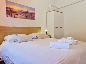 Un lit bien fait, avec des oreillers, une lampe de chevet, une armoire et un tableau représentant une scène de plage.