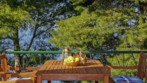 Una mesa de madera con un farol y una cesta de frutas en un balcón con vistas al agua, con sillas y cojines.