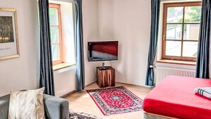 A room with two windows, a flat-screen TV, a radiator, a red bed, a grey sofa, and a patterned rug. - Dr.Steiner by Interhome (Zell am See)