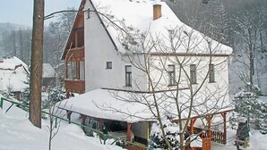 A snow-covered house with a large balcony, surrounded by trees and other buildings.