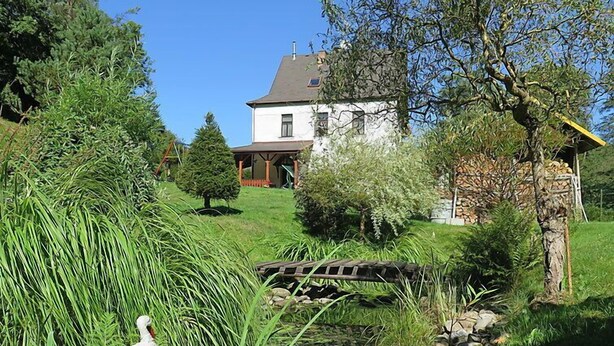 A white house with a stone bridge, surrounded by lush greenery and a pond.