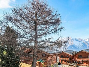 Una cabaña de madera con un gran árbol en primer plano, una montaña al fondo y un cielo despejado.