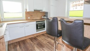 Eine moderne Küche mit weißen Schränken, einer Holzausziehtafel und einem eingebauten Backofen. Zwei schwarze Barhocker und ein Fenster mit Blick auf ein Gebäude sind vorhanden.
