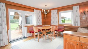 Ein gemütlicher Raum mit einem Holzespeisetisch, Stühlen und einer Bank. Es gibt Fenster mit Vorhängen, eine Pendelleuchte und einen Holzkasten.