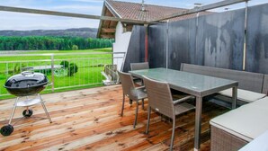 Ein Balkon mit Holzboden, einem Tisch mit Stühlen, einem Grill und Blick auf grüne Felder und Berge.