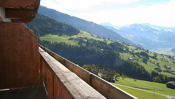 Un balcon en bois donnant sur un paysage montagneux, avec des maisons et des champs verdoyants.