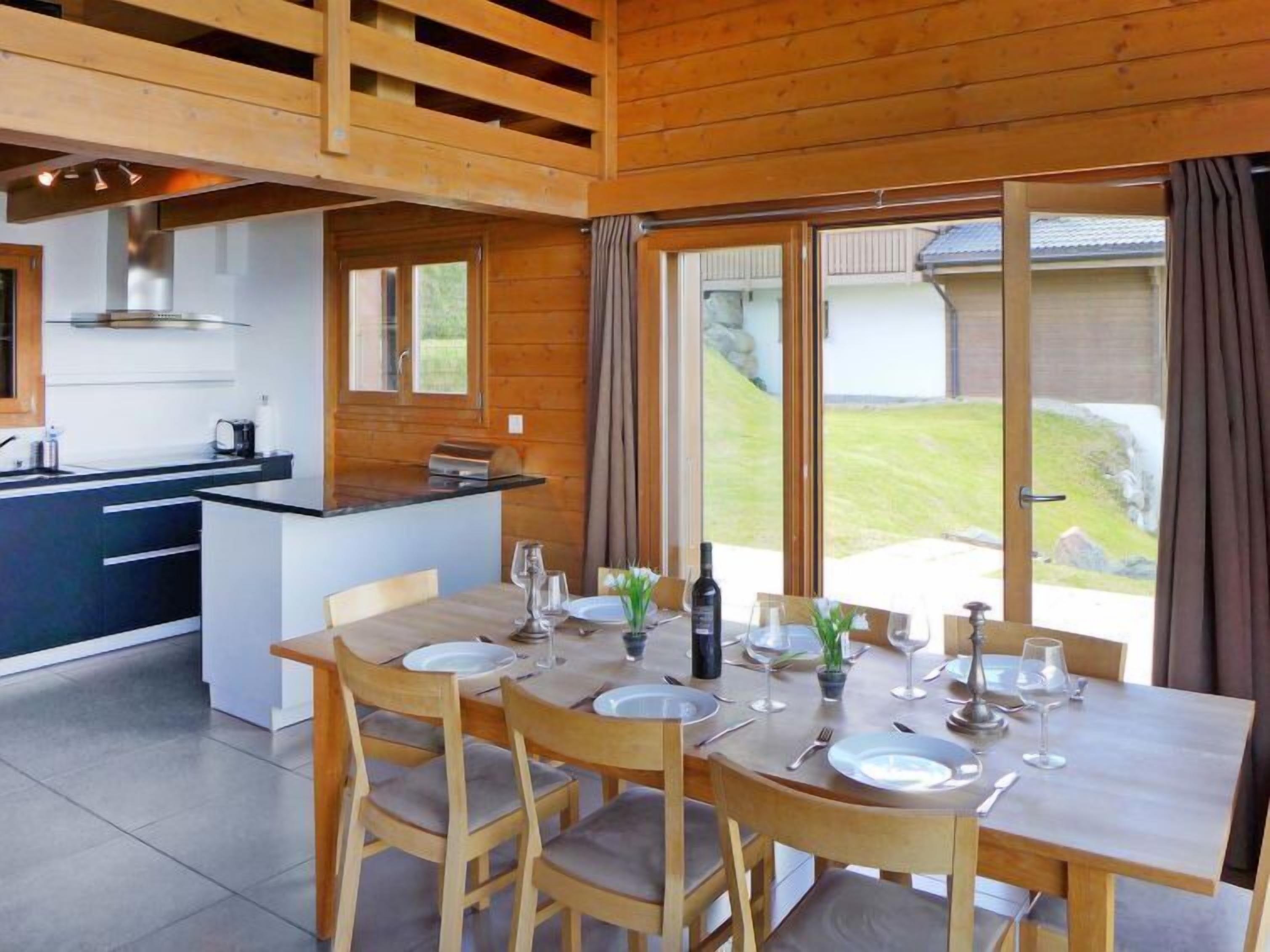 Une cuisine moderne dotée d’un îlot central, d’une table à manger en bois avec des chaises, et de grandes fenêtres offrant une vue sur l’extérieur.