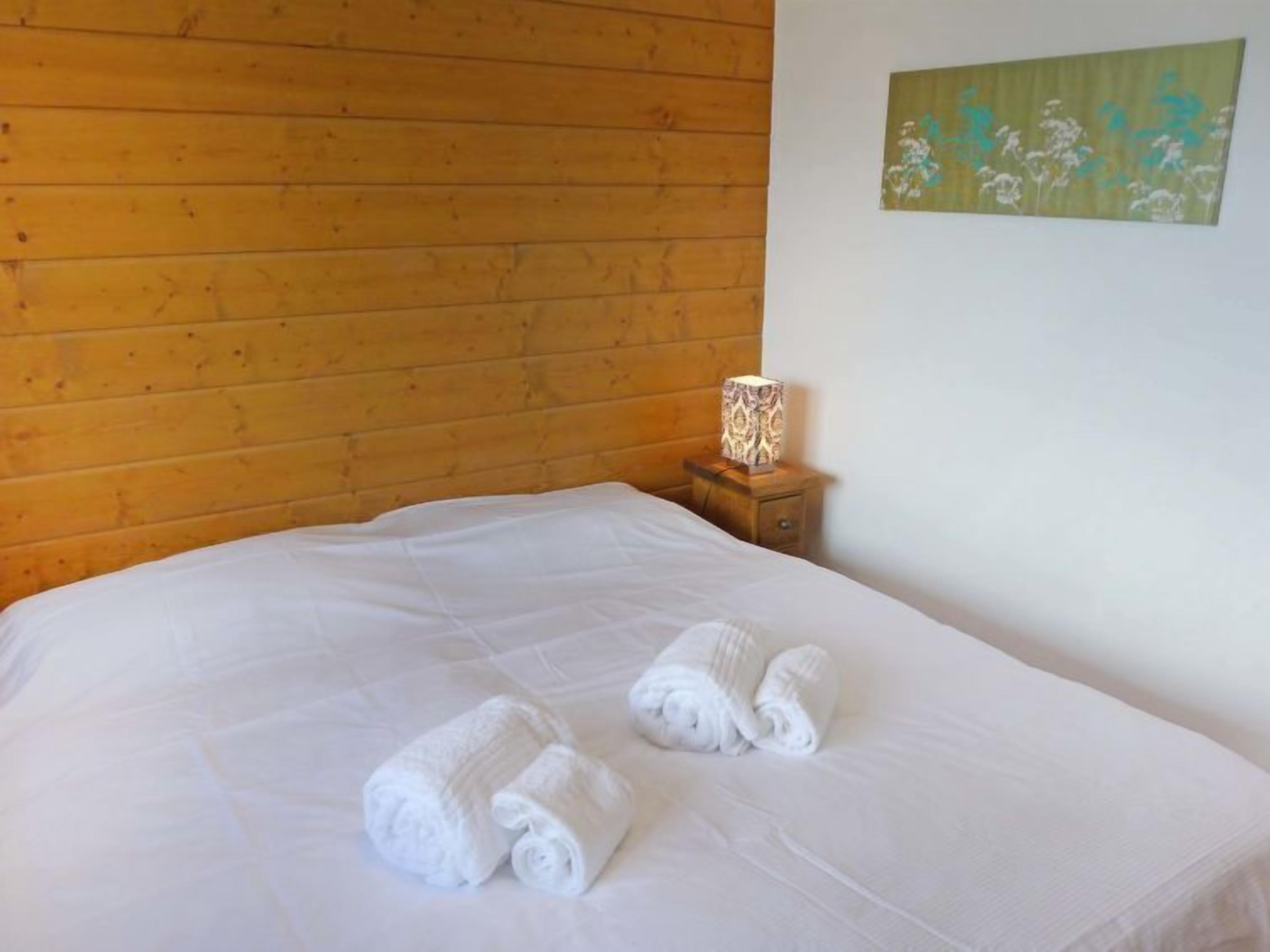 Un lit bien fait, avec des draps blancs et deux serviettes enroulées, une tête de lit en bois, une table de chevet avec une lampe et un tableau floral accroché au mur.