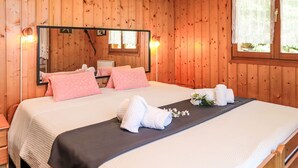 Ein Schlafzimmer in einer Holzhütte mit einem Bett, Kissen, einem Nachttisch, einem Spiegel und einem Fenster mit Spitzenvorhängen.