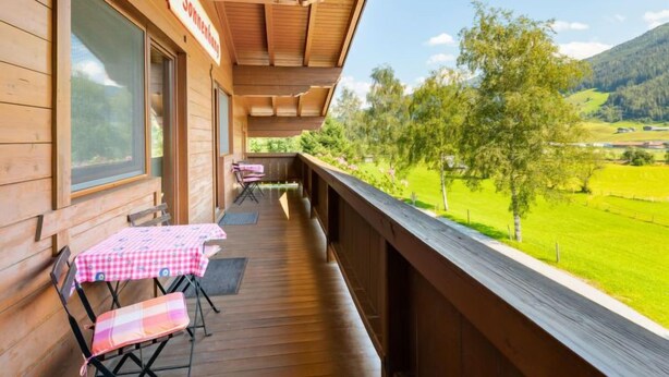 Ein Holzbalkon mit Tisch und Stühlen, der auf eine grüne Landschaft und Berge blickt.