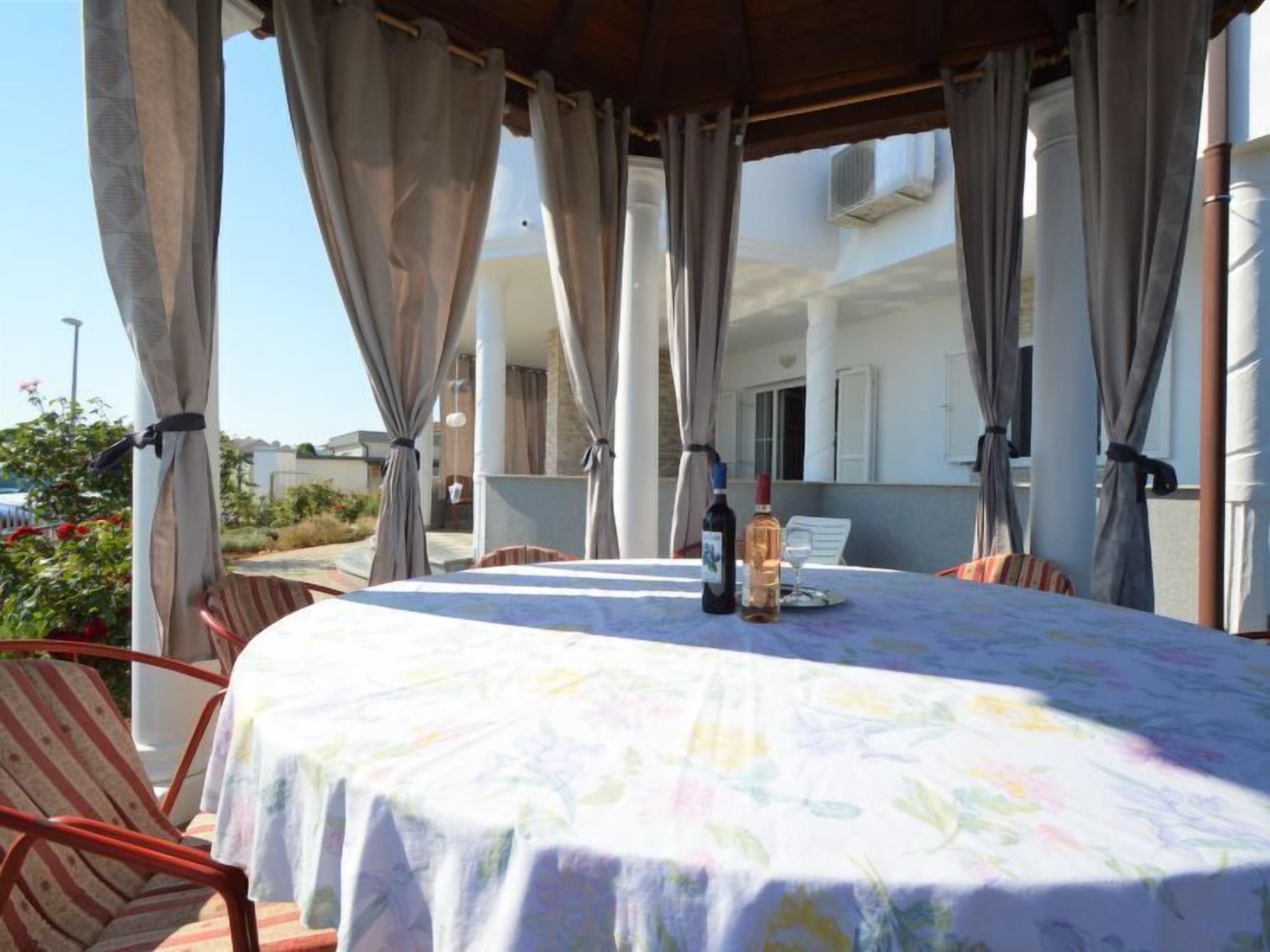 Un espace extérieur abrité, aménagé pour les repas, avec une table dressée pour deux personnes, ornée d'une nappe à motifs floraux et de deux bouteilles de vin.