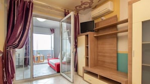 Una habitación con puerta de vidrio corredera que da a un balcón, una cama roja, un armario de madera y un televisor en la pared.