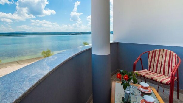 Un balcón con vista a un lago tranquilo y montañas a lo lejos, una silla a rayas rojas y una mesa con un florero y un vaso.