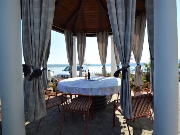 A covered outdoor seating area with a round table and chairs, overlooking a body of water.