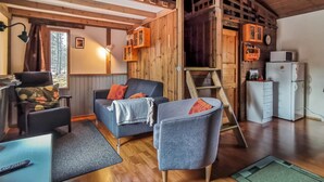 Un intérieur de chalet chaleureux avec une chambre en mezzanine, des murs en bois, un canapé, un fauteuil, un bureau et un réfrigérateur.