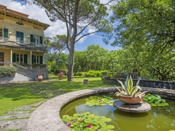 Un jardín con un estanque, un edificio con balcón y un árbol grande.