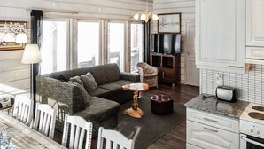 A modern living room with a grey sofa, a small round table, and a white chair. There is a television on a wooden cabinet, a kitchen area with white cabinets, and a window with blinds. - Kerkänperä by Interhome (Sodankylä)