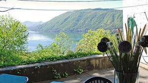 Una terraza con vista a un lago y montañas, un jarrón con flores y mobiliario exterior.