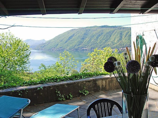 Una terraza con vista a un lago y montañas, un jarrón con flores y mobiliario exterior.