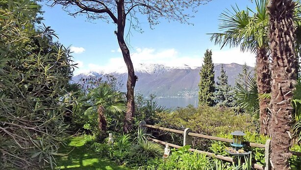 Un giardino con un recinto in legno, diverse piante, una piccola statua e vista sulle montagne e un lago.