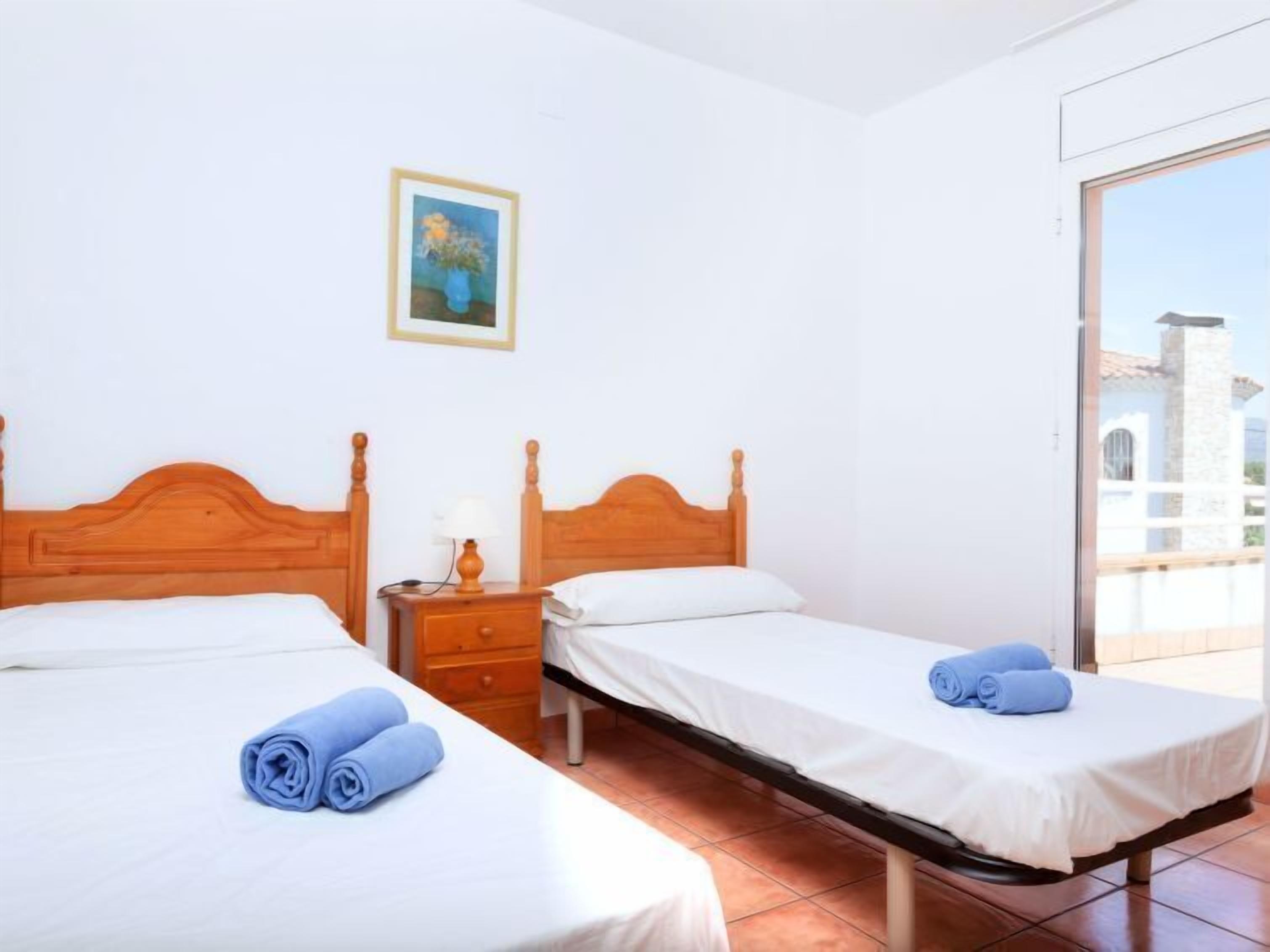A hotel room with two wooden beds, white linens, a small bedside table with a lamp, and a painting on the wall.