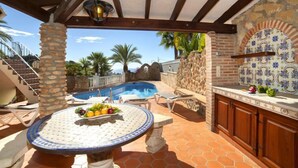 A poolside area with a stone pillar, a round table with fruit, lounge chairs, and a tiled backsplash. - Roberto by Interhome (Benissa)
