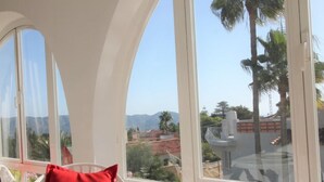 Una habitación iluminada por el sol con vistas a palmeras y montañas, una mesa redonda con un cuenco y cojines rojos.