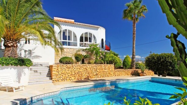 Un área de piscina con un muro de piedra, palmeras y un edificio con ventanas arqueadas.