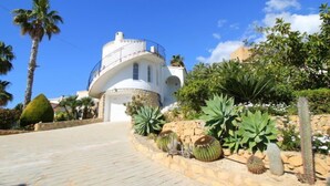 Un edificio de techumbre blanca y curva con balcón, rodeado de plantas tropicales y un sendero de piedra.