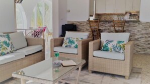 Sala de estar con muebles de mimbre, una mesa de centro de vidrio y una cocina con salpicadero de piedra.