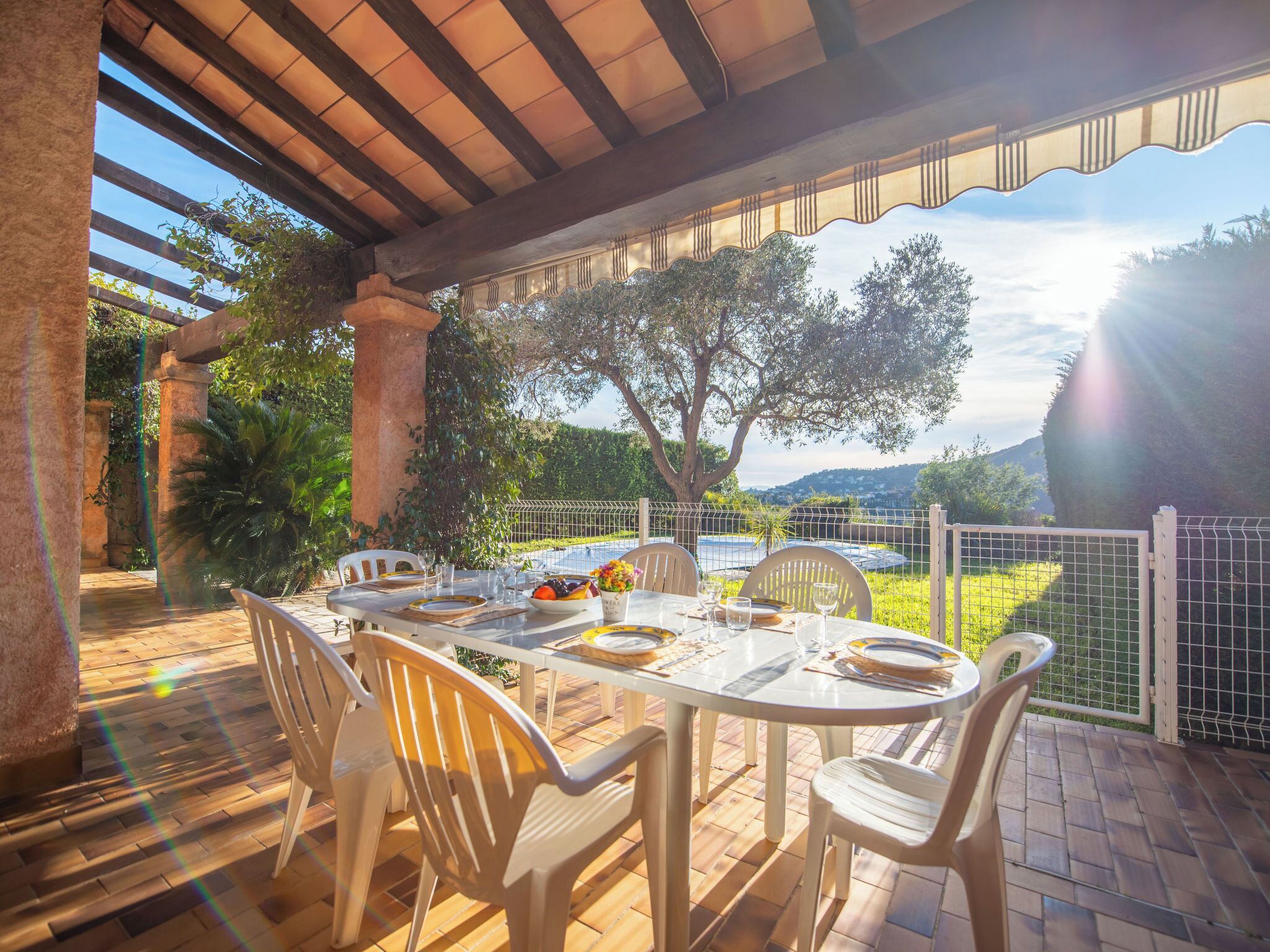 An outdoor dining area with a table set for four, surrounded by chairs, under a pergola with a view of a garden and distant hills.