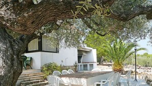 An outdoor dining area with a stone table and white plastic chairs, surrounded by trees and a white building with a balcony. - El Rinconcito by Interhome (L'Ampolla)