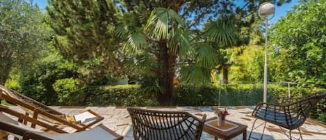 A patio area with wicker chairs and a table, surrounded by lush greenery and a clear blue sky.