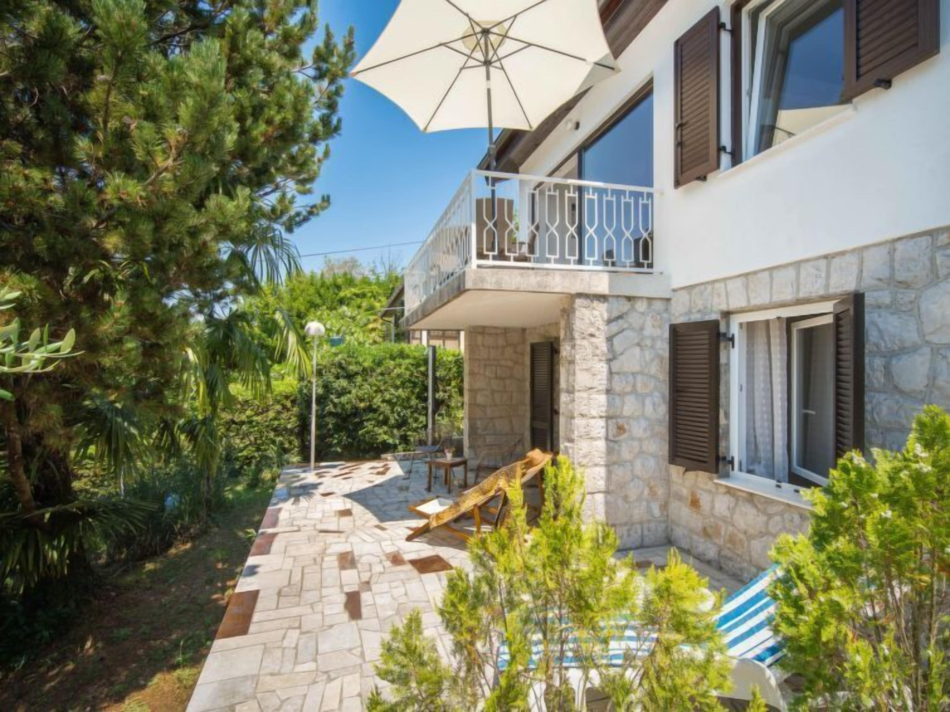Una casa de dos pisos con fachada de piedra, un balcón con barandilla metálica y un patio con sombrilla blanca.